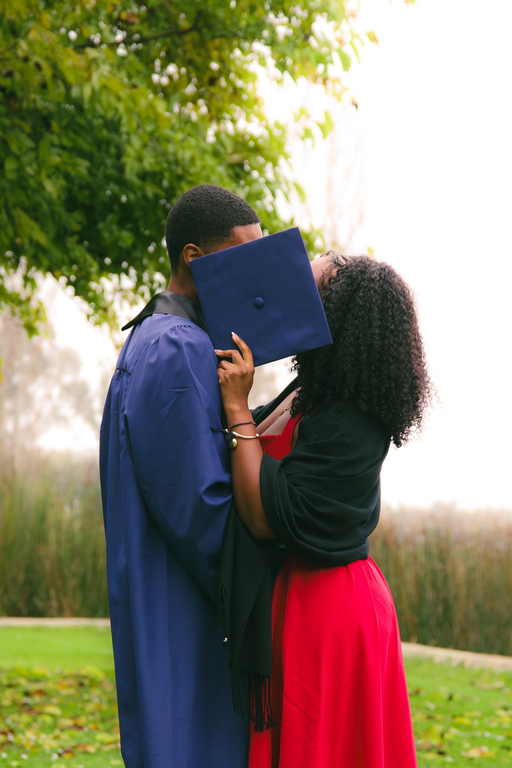 A couple shares a kiss hidden behind a blue graduation cap in a park.