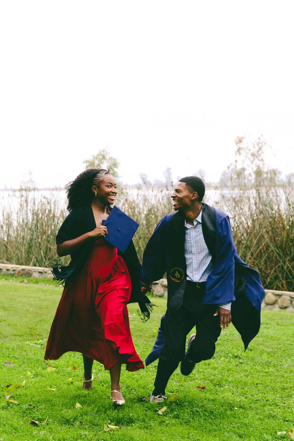 A joyful couple runs together outdoors, the man wearing a blue graduation gown.