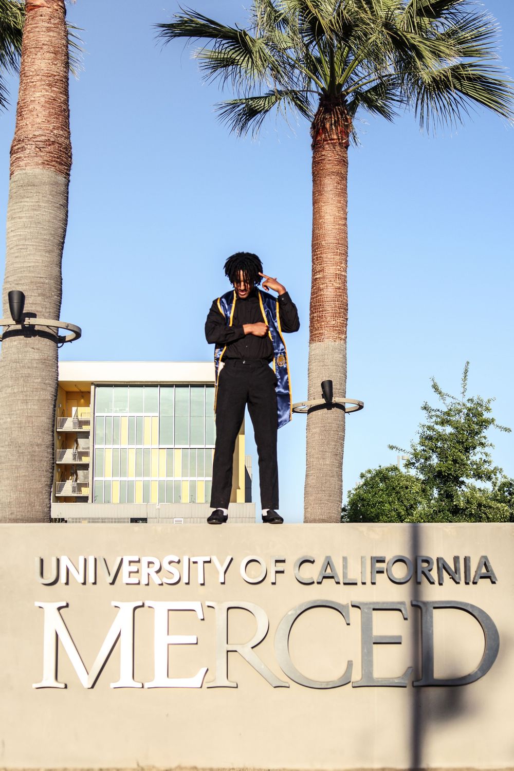 Graduate stands atop University of California Merced campus sign between tall palm trees at dusk.