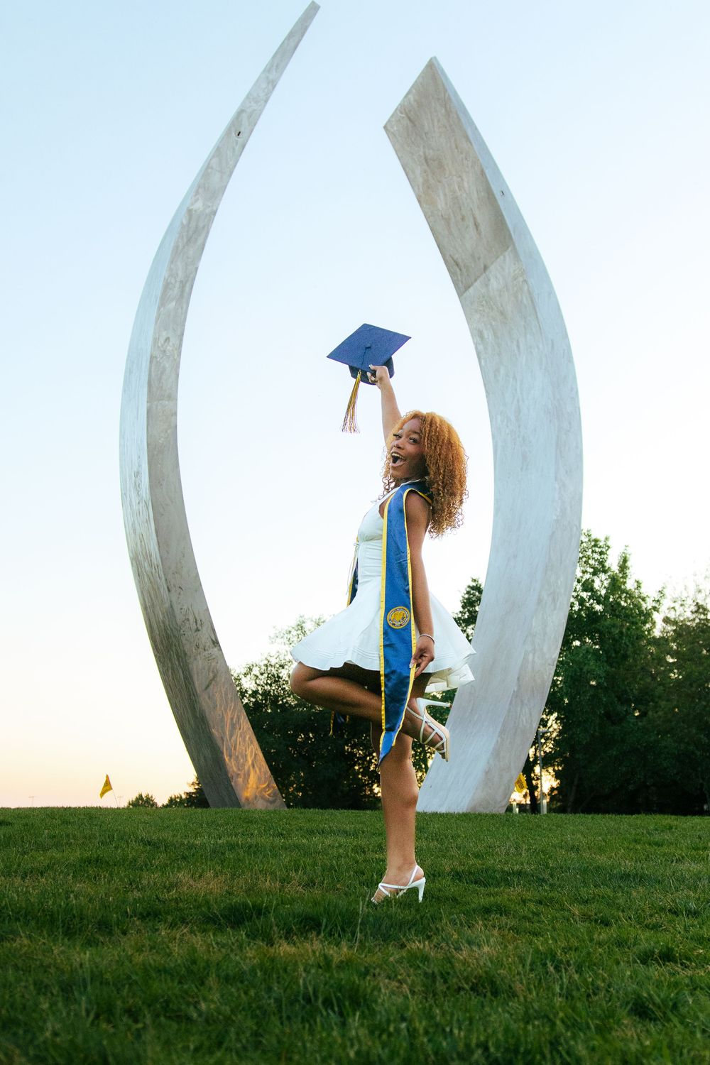 Joyful graduate celebrates outdoors holding cap aloft near large curved sculpture on green campus lawn.