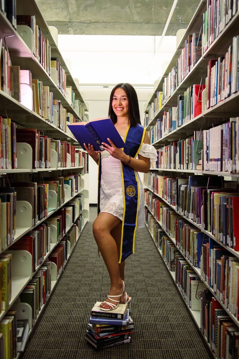 Smiling graduate in white dress with blue sash standing on books in library.