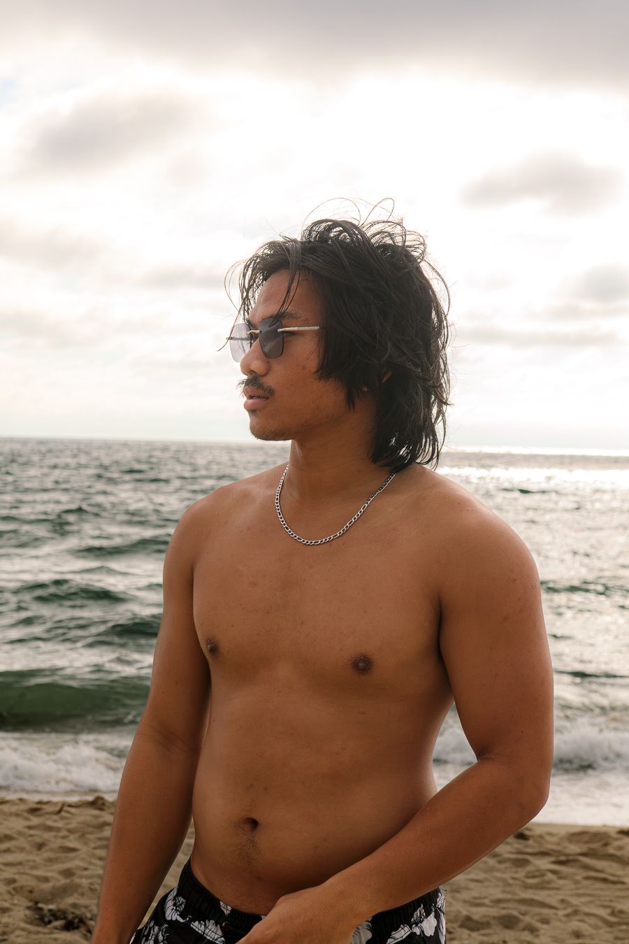 Shirtless young man with sunglasses and silver chain stands confidently on sandy beach by ocean.