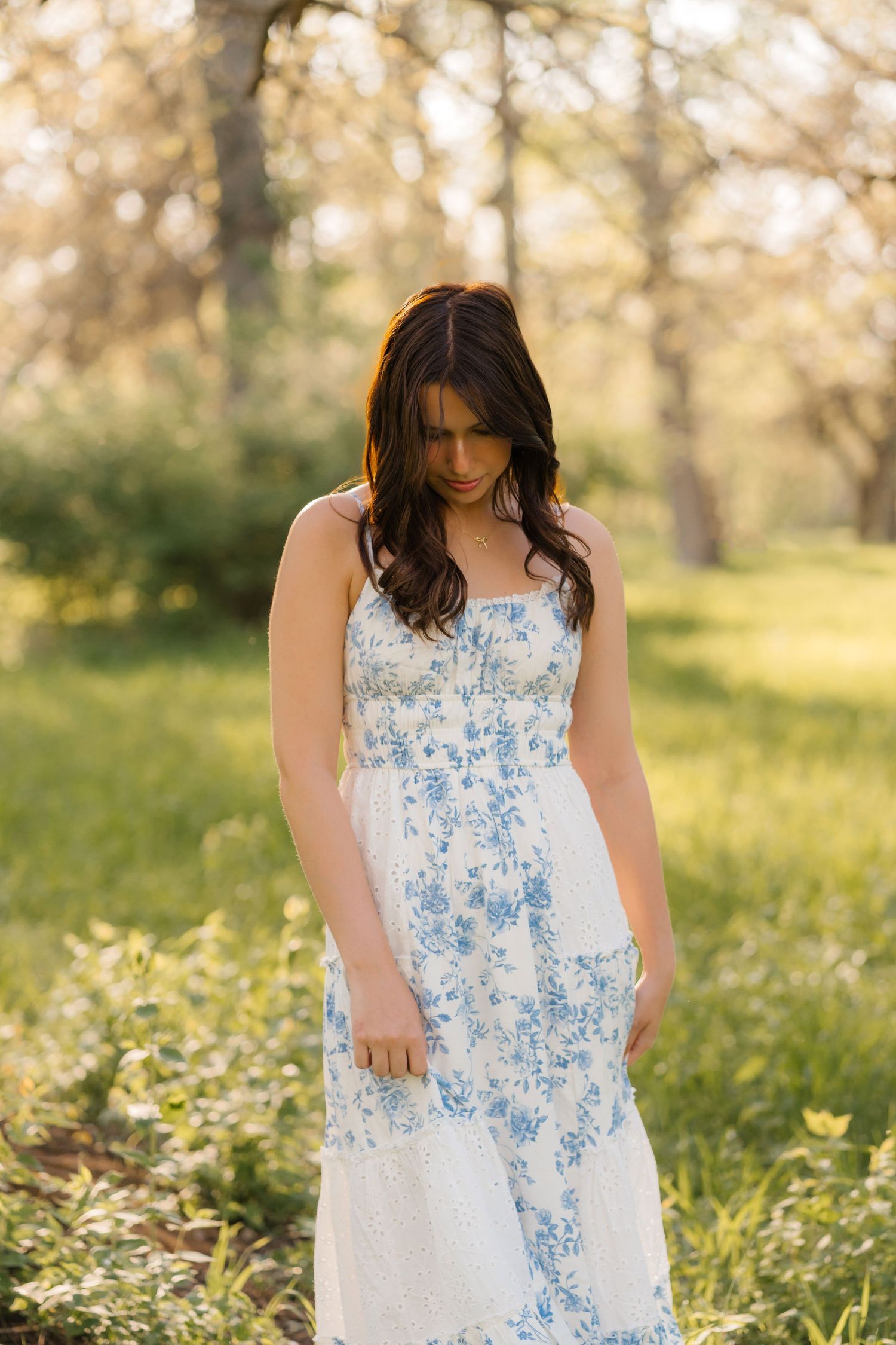 A person wearing a white and blue floral maxi dress poses in a sunlit forest clearing during golden hour.