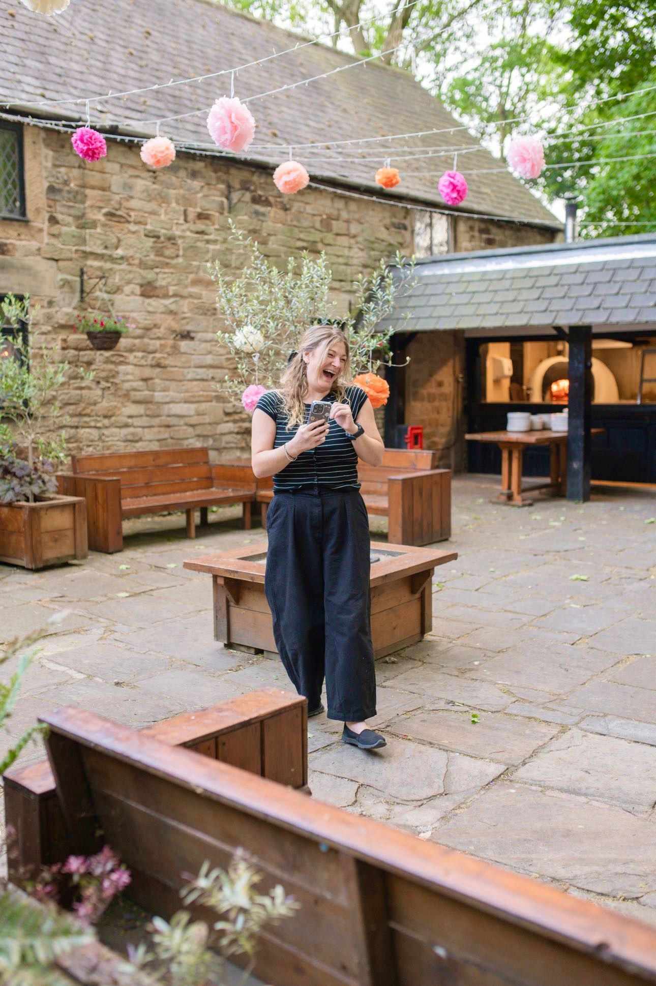 Emmaweekesart content creator stood with phone in hand at a wedding laughing.