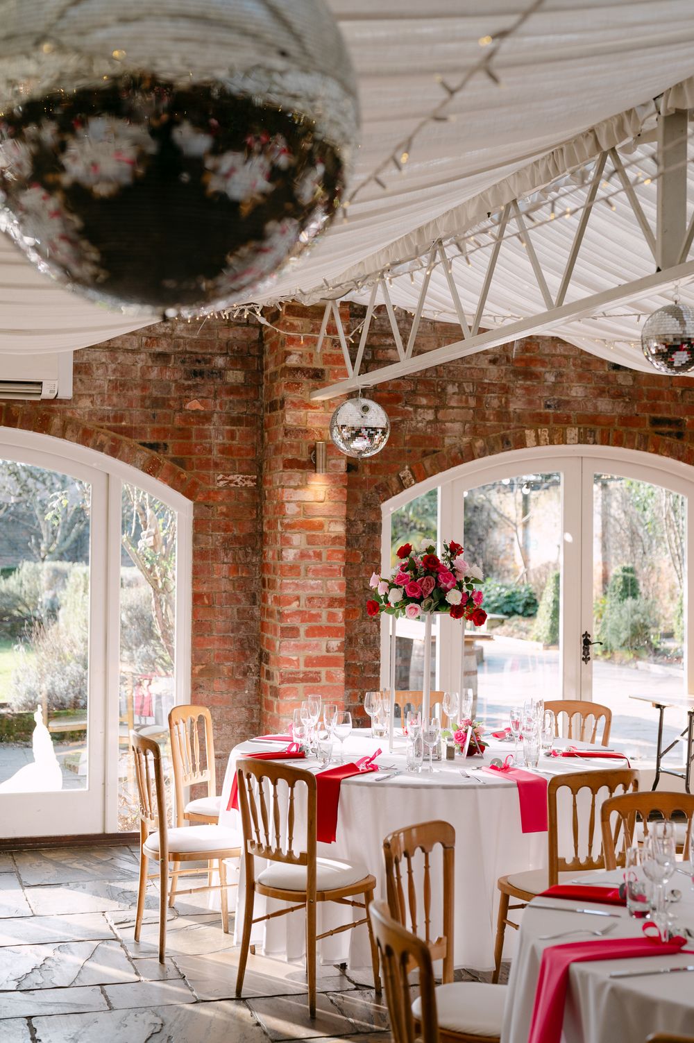 Rustic indoor wedding venue with exposed brick walls, white tables, wooden chairs and red accent decor.