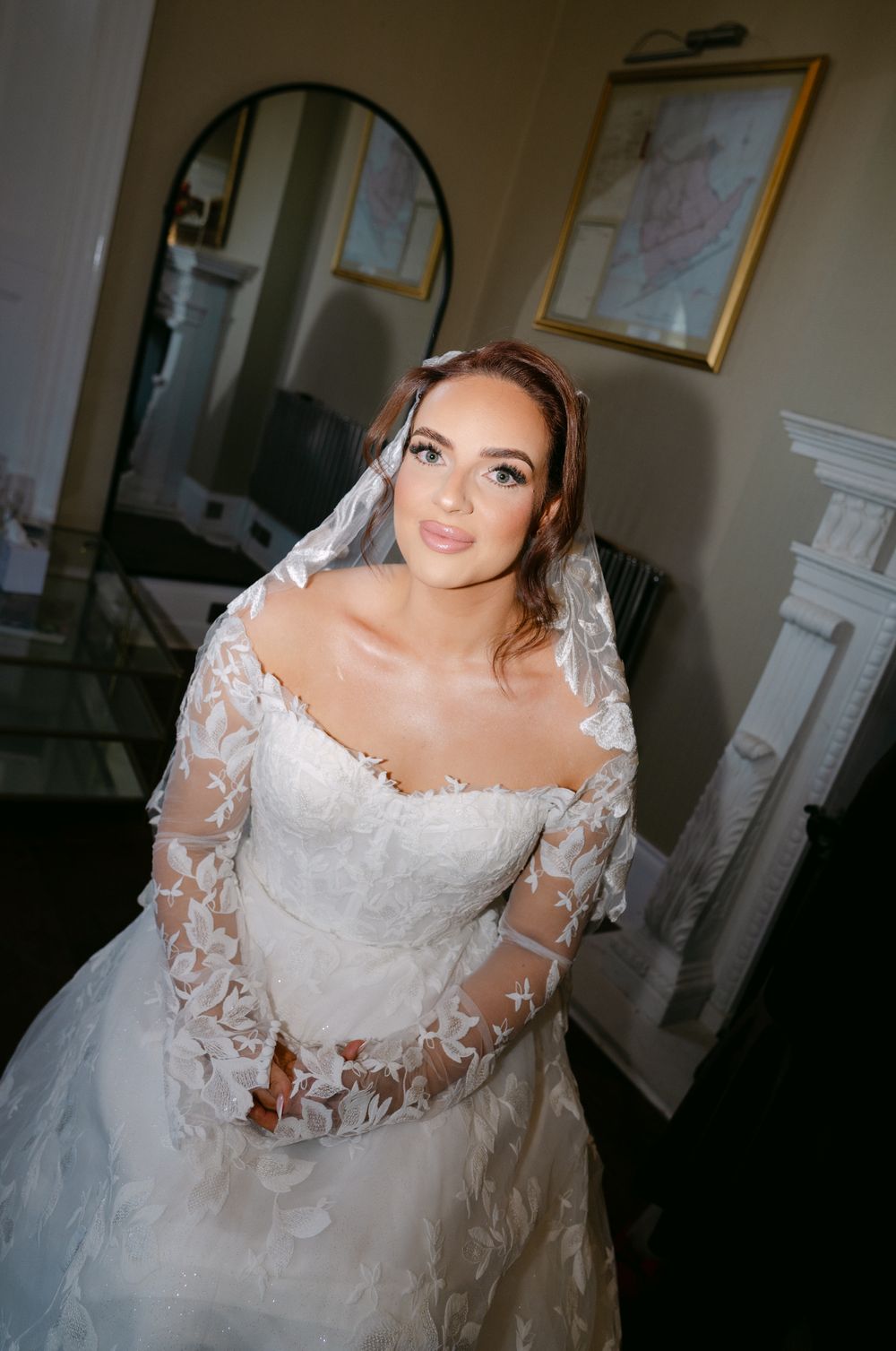 A bride in a long-sleeved lace wedding gown with sheer details poses indoors near a framed artwork.