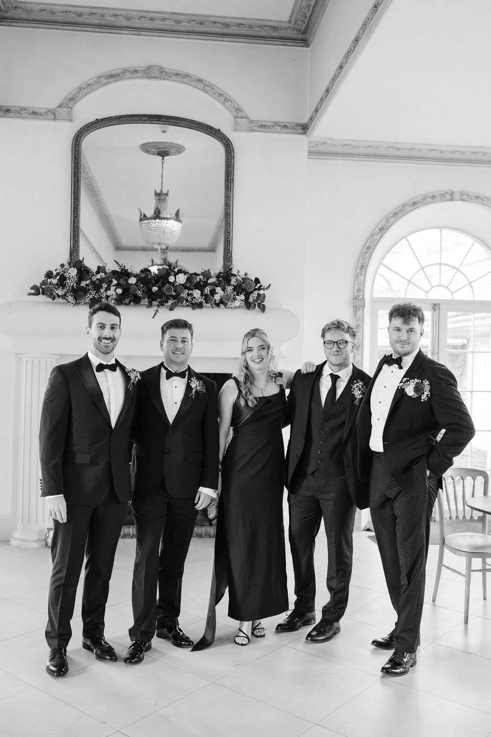 A group of groomsmen in black tuxedos and bow ties pose together in an elegant white room with ornate moldings.