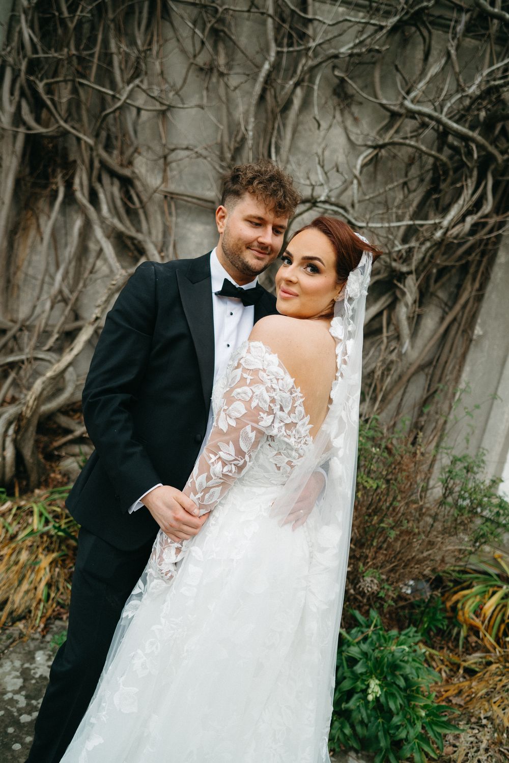 Newlywed couple shares intimate moments in a garden setting with bare branched trees and greenery.
