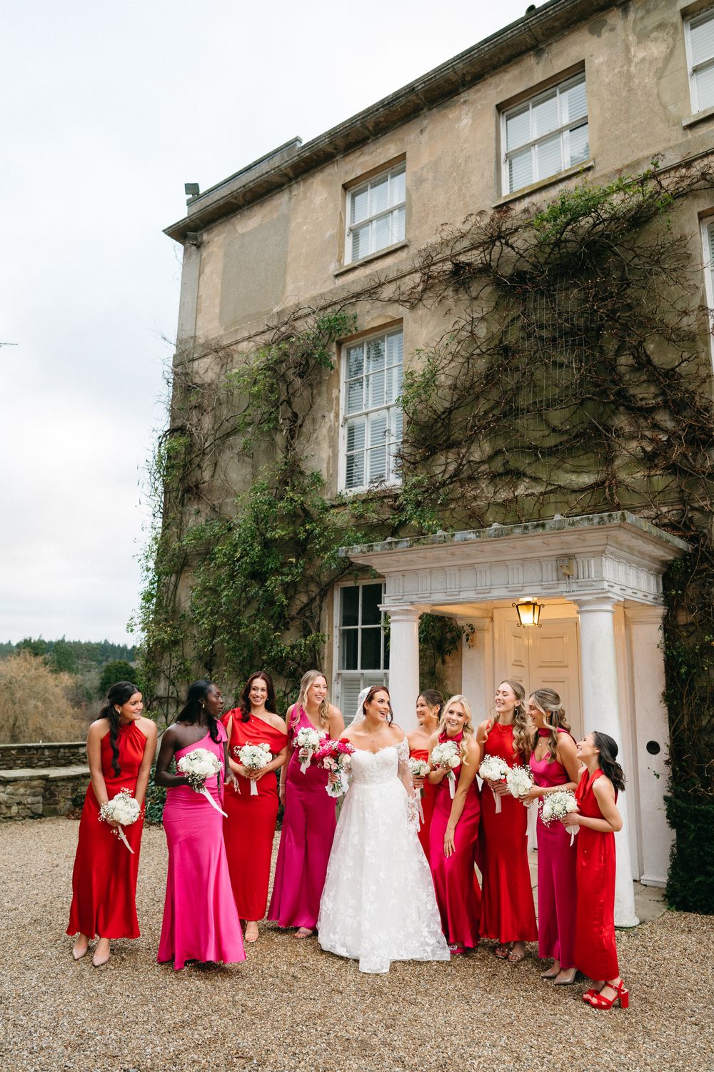 A bridal party wearing red and pink dresses poses outside an ivy-covered stone mansion with elegant columns.