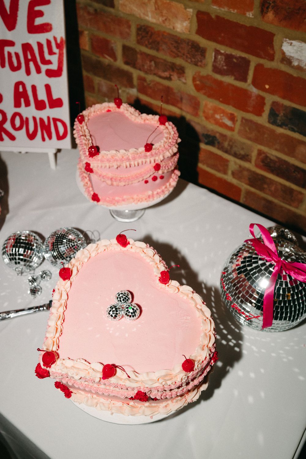 Pink heart-shaped cake with red decorative cherries and silver disco balls on white surface against brick wall.
