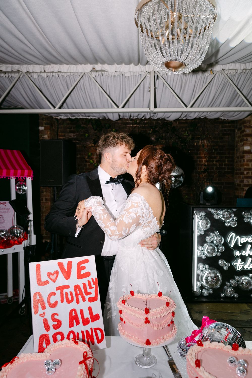 A couple dances at a wedding reception with a decorated cake table featuring a 'Love Actually' themed sign.