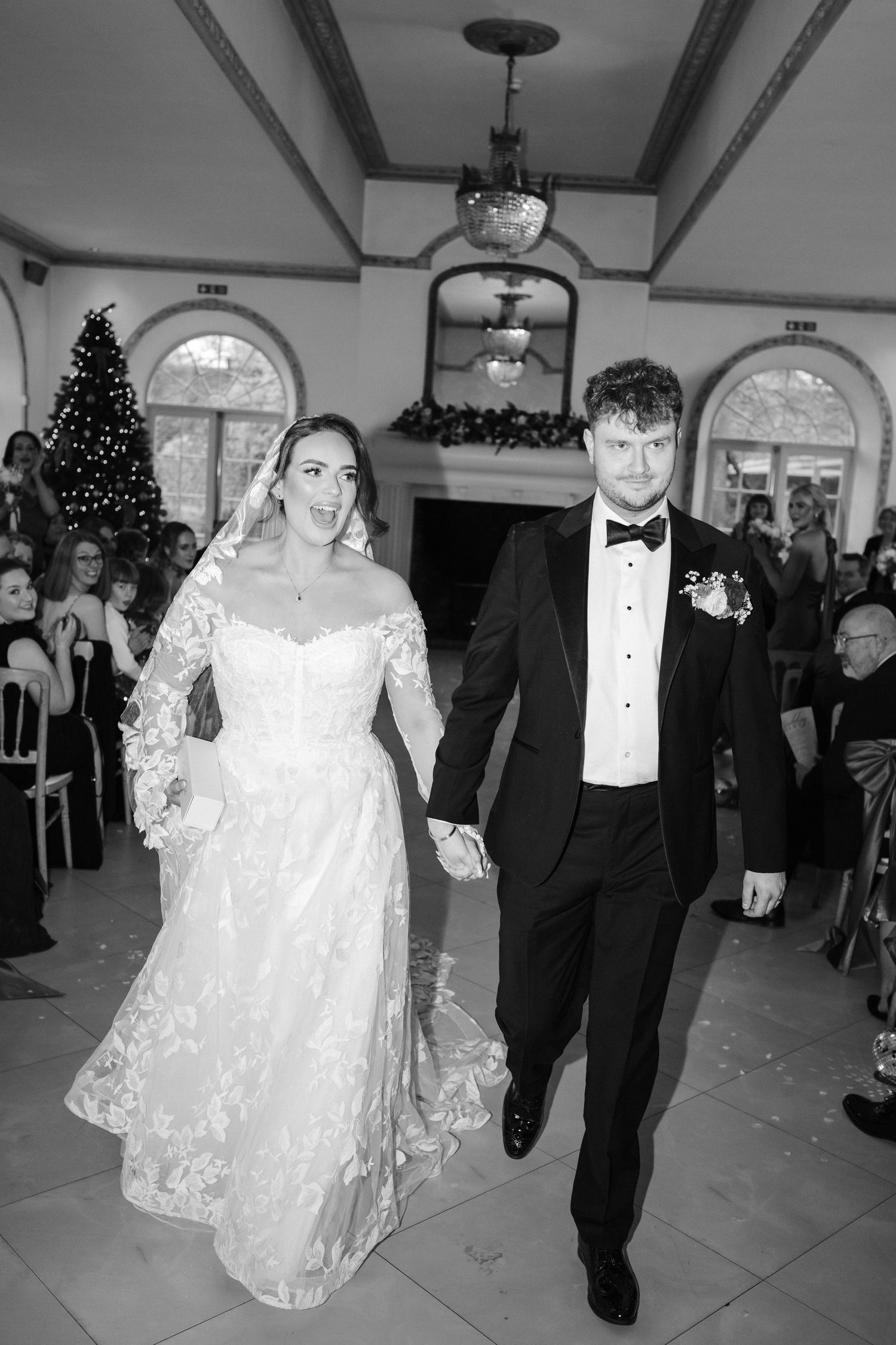 A bride and groom walk down a reception hall aisle decorated with Christmas trees and holiday decor in black and white.