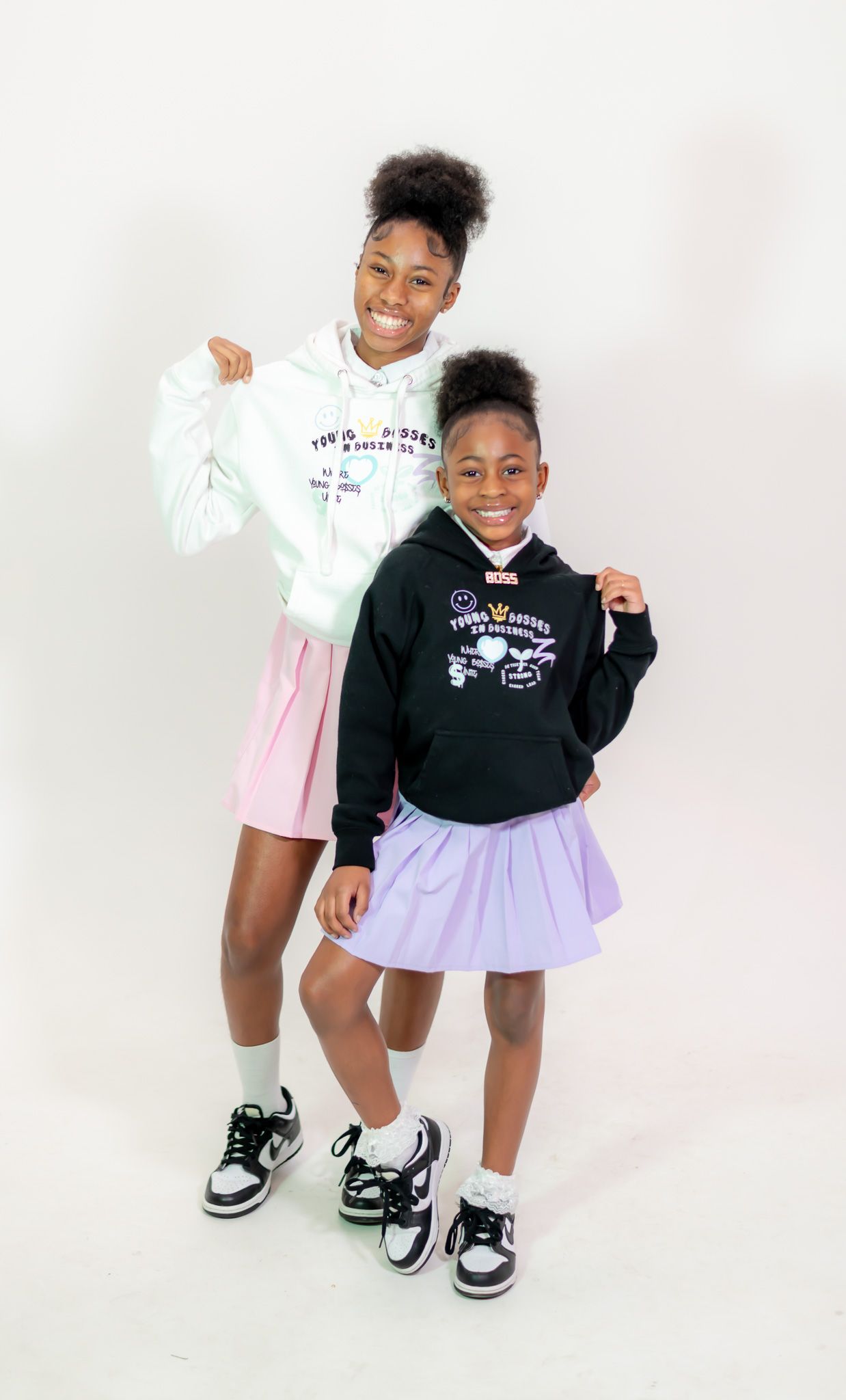 Two people wearing contrasting black and white hoodies with pink and purple skirts and matching sneakers pose against white background.