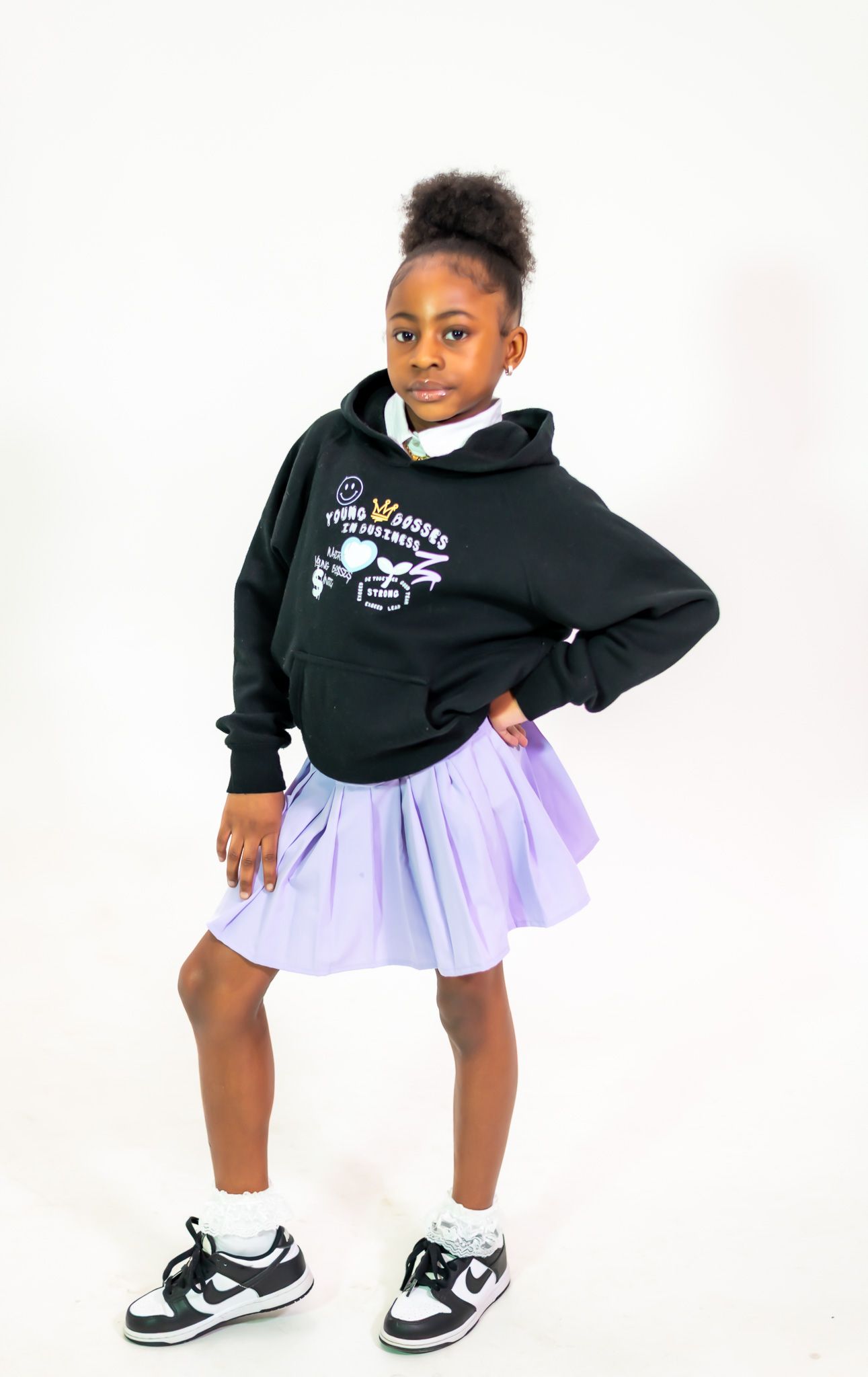 A young model poses in a black hoodie, lavender tulle skirt and sneakers against a white background.