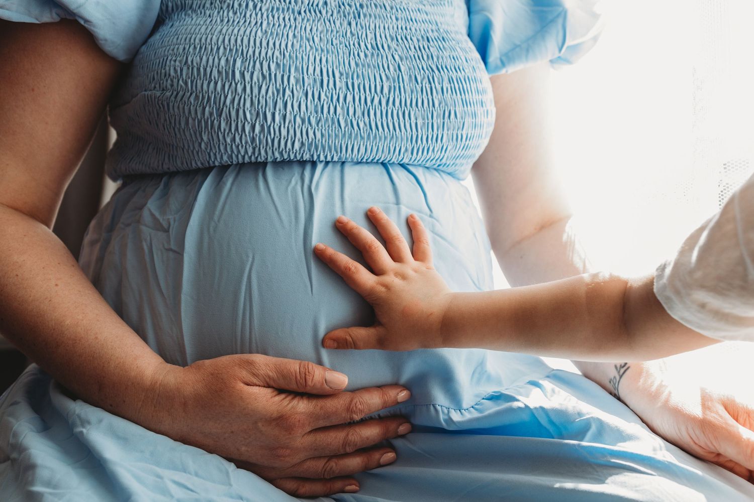A pregnant woman's belly is cradled by gentle hands while wearing light blue clothing in soft natural lighting.