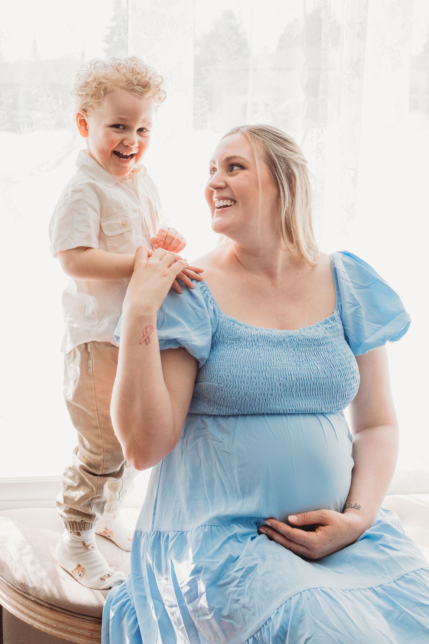 A pregnant woman in a light blue dress shares a joyful moment with a young child standing on furniture.