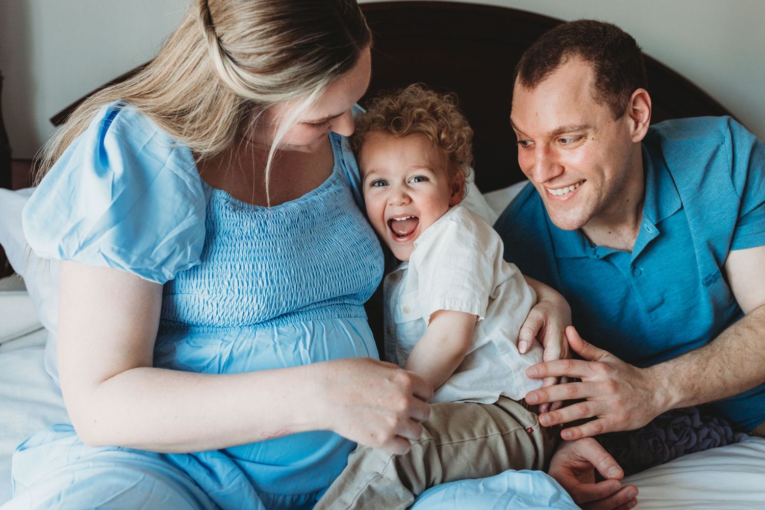 A family shares a joyful moment together while lounging on a bed, with parents wearing blue and a laughing child between them.