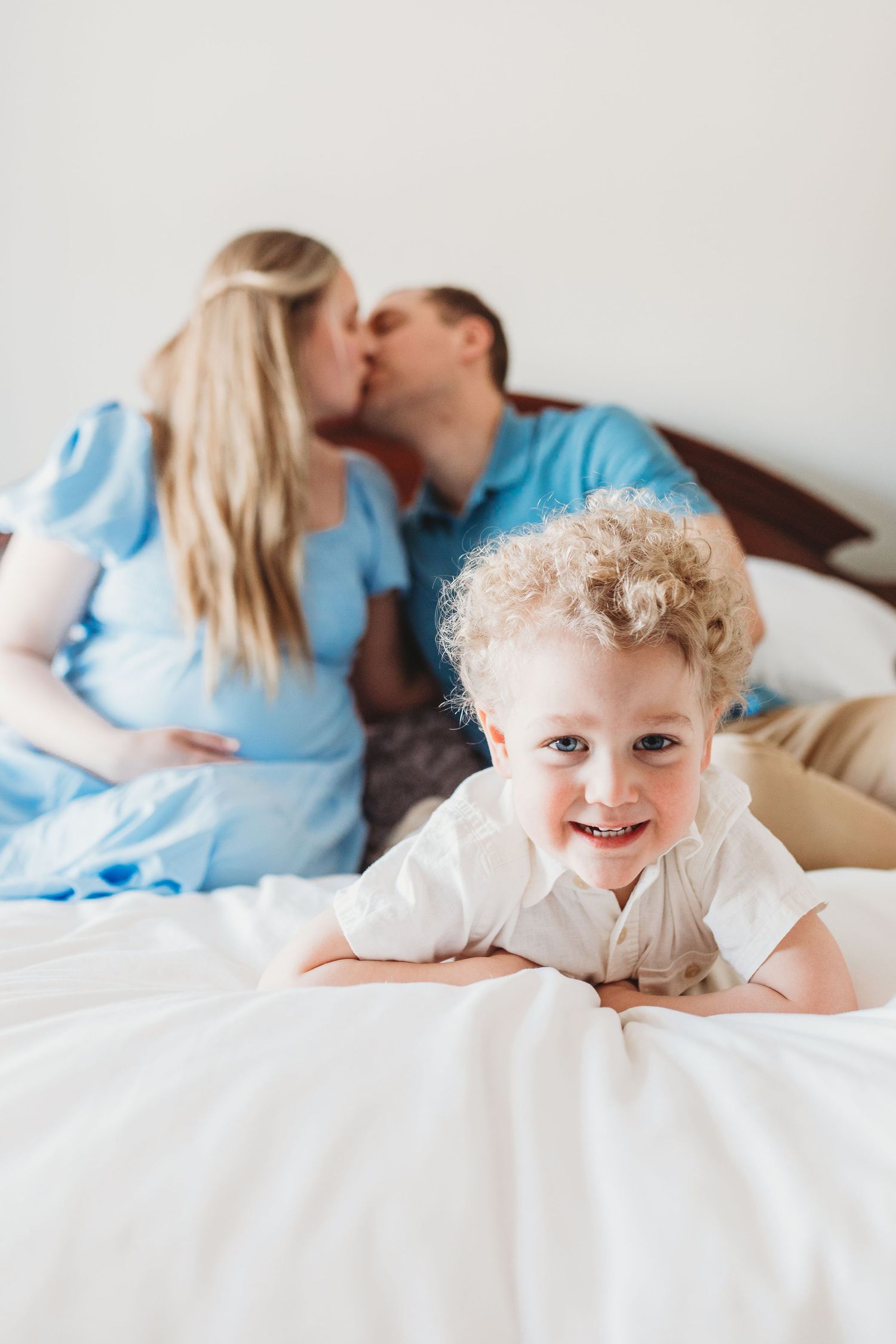 A happy toddler with curly blonde hair lying on a white bed while a couple embraces in the background.