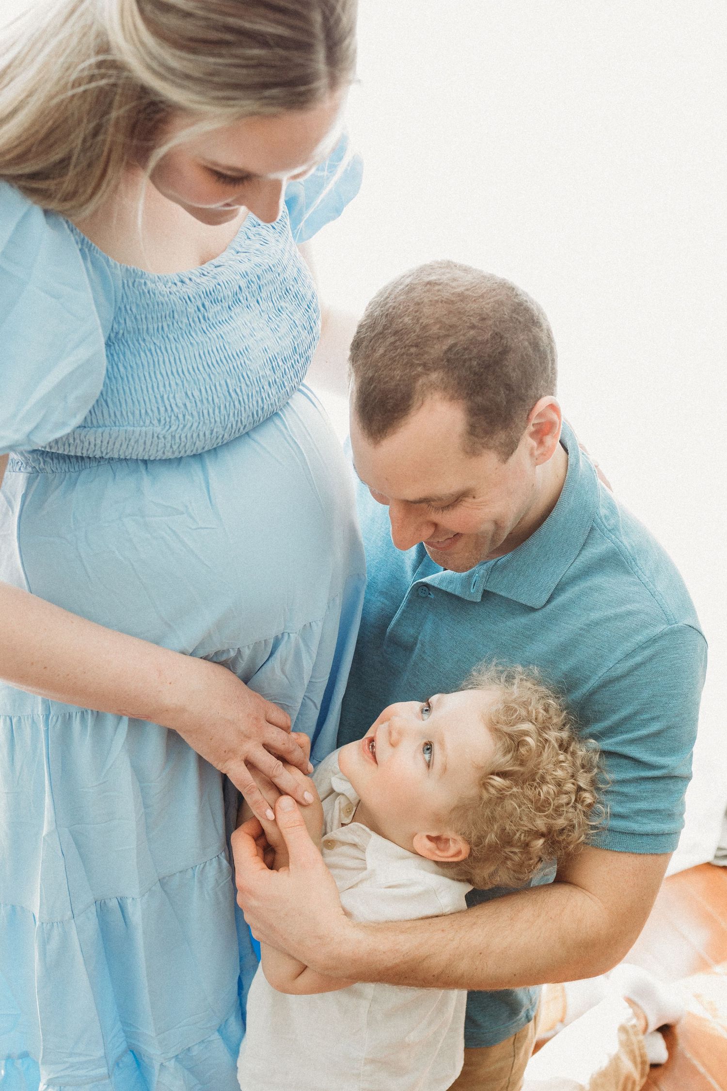 A pregnant woman in a blue dress and partner share a tender moment with their toddler with curly hair.