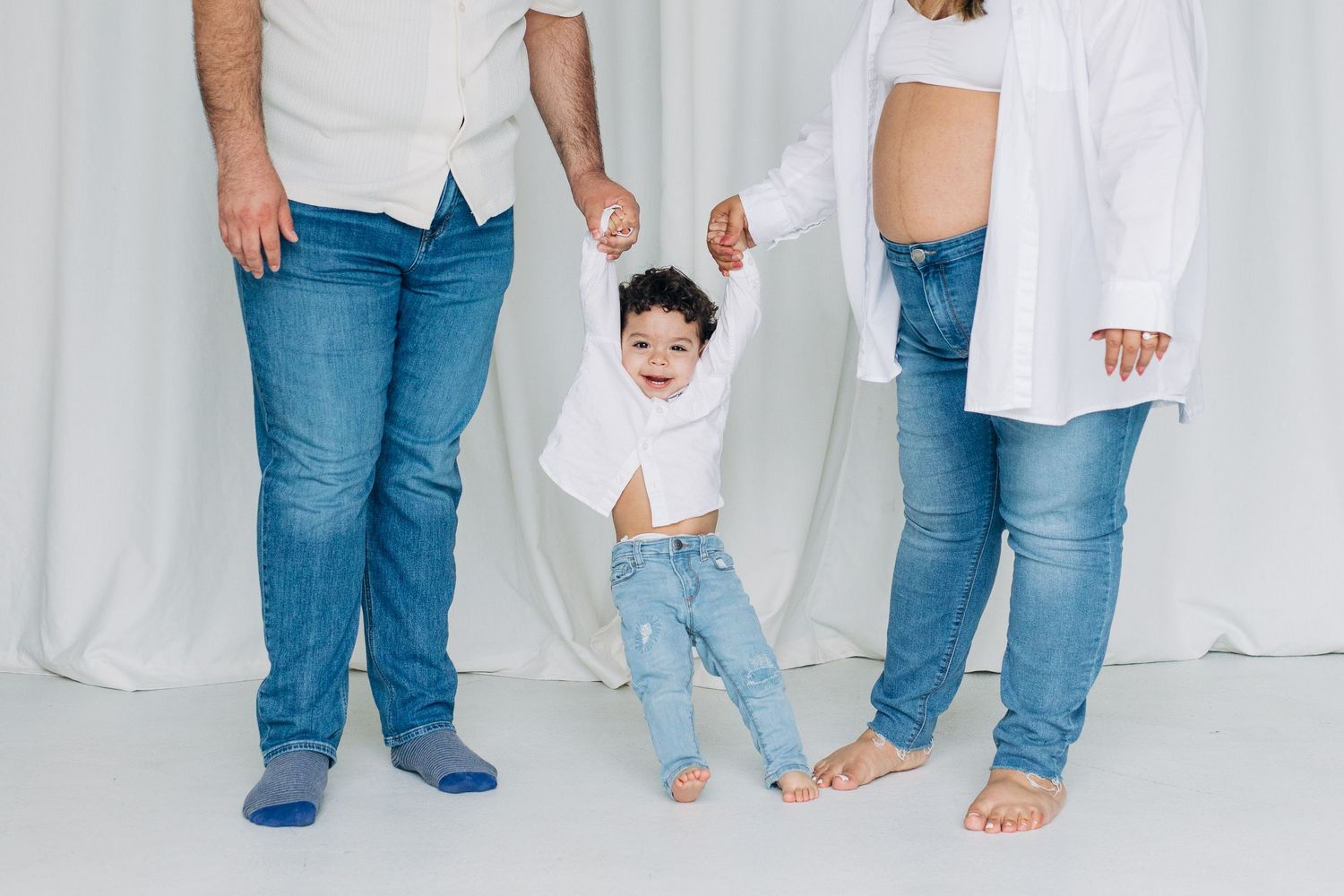 A group of people wearing blue jeans and white shirts stand together showing only their legs and feet against a white backdrop.