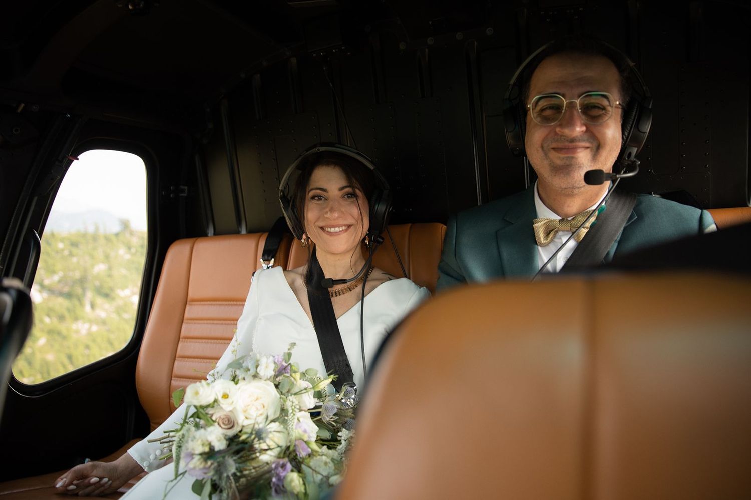 Smiling bride in white dress holding bouquet sits beside groom wearing headset inside helicopter.