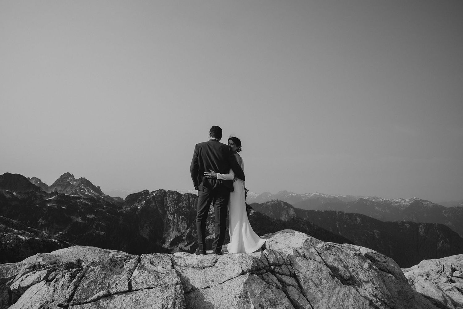 Black and white photo of bride and groom standing on rocky summit overlooking mountain range.
