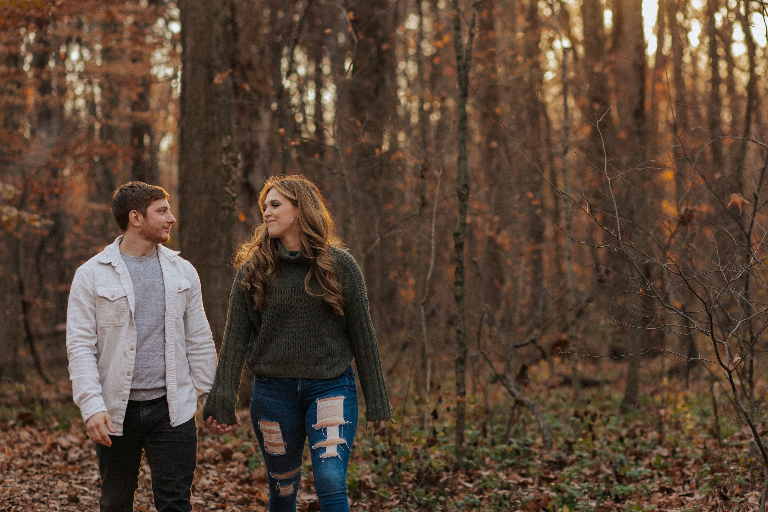 Couple walking through autumn forest at sunset with golden light filtering through bare trees.