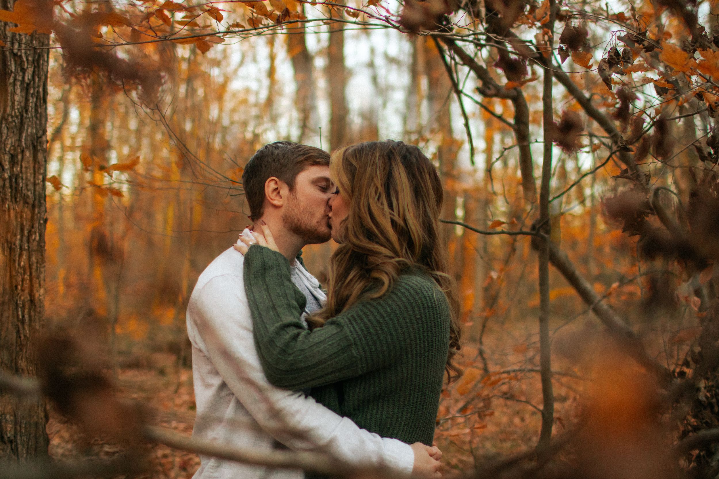 A romantic couple shares a tender kiss in an autumn forest surrounded by warm orange foliage.
