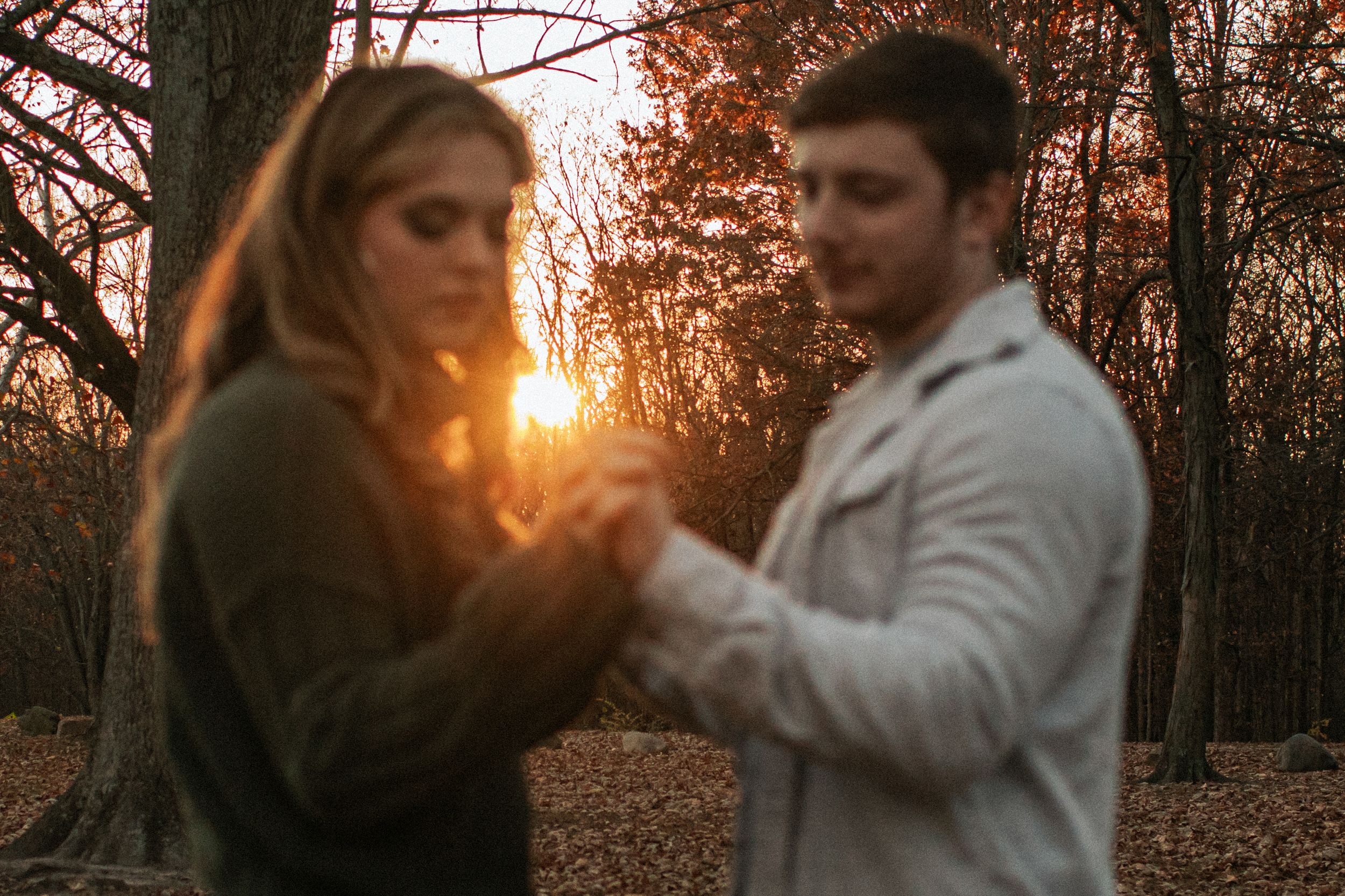Two people hold hands in a romantic pose during golden hour sunset in an autumn forest setting.