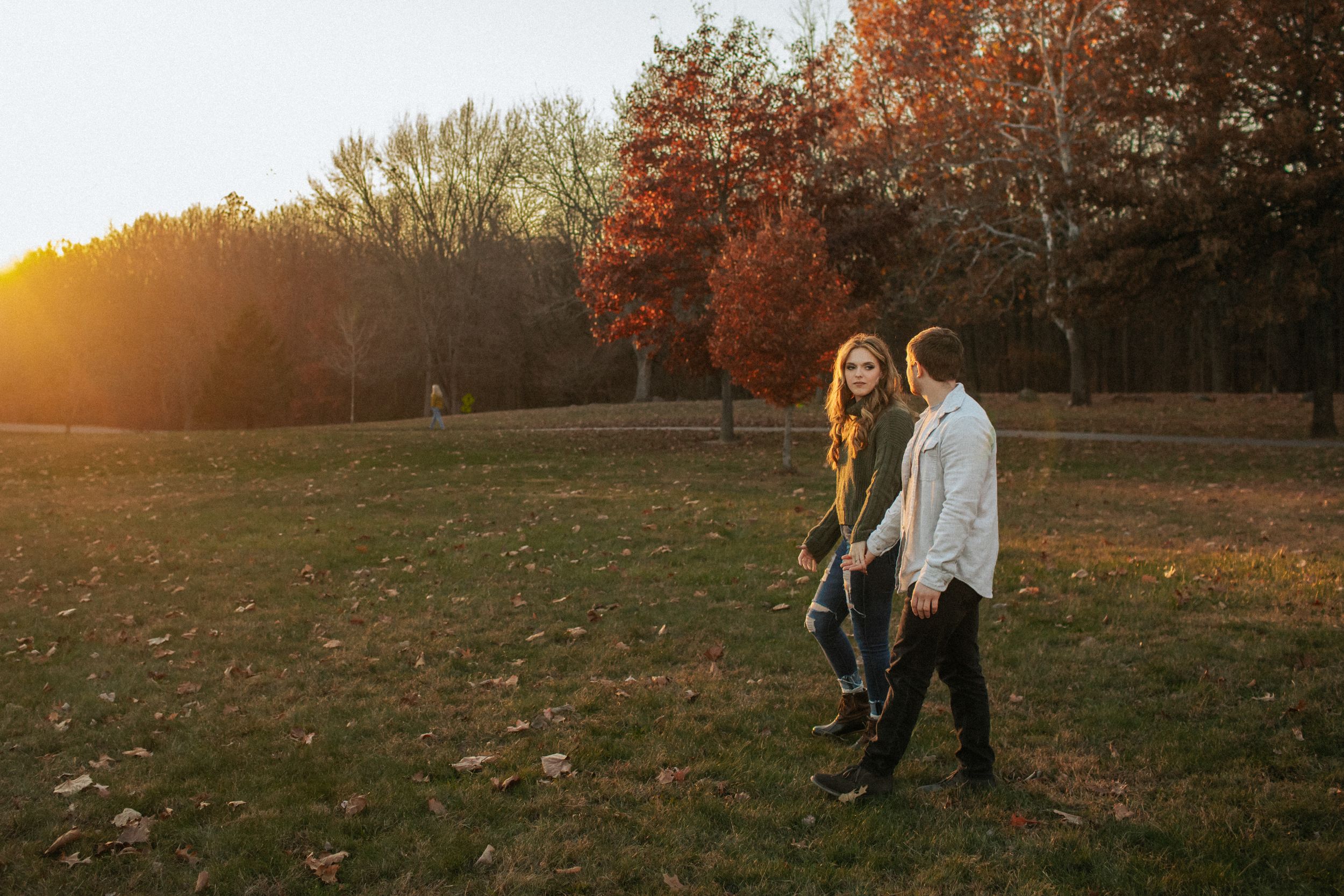 A couple walks together through a golden autumn field at sunset with trees in the background.