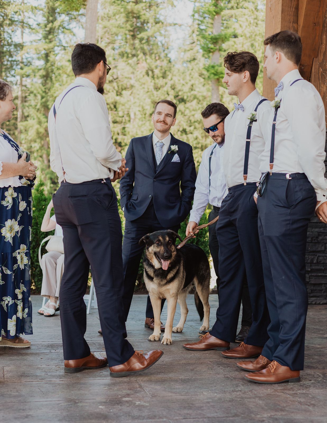 Groom and groomsmen in navy suits with suspenders gathered with a German Shepherd dog outdoors.