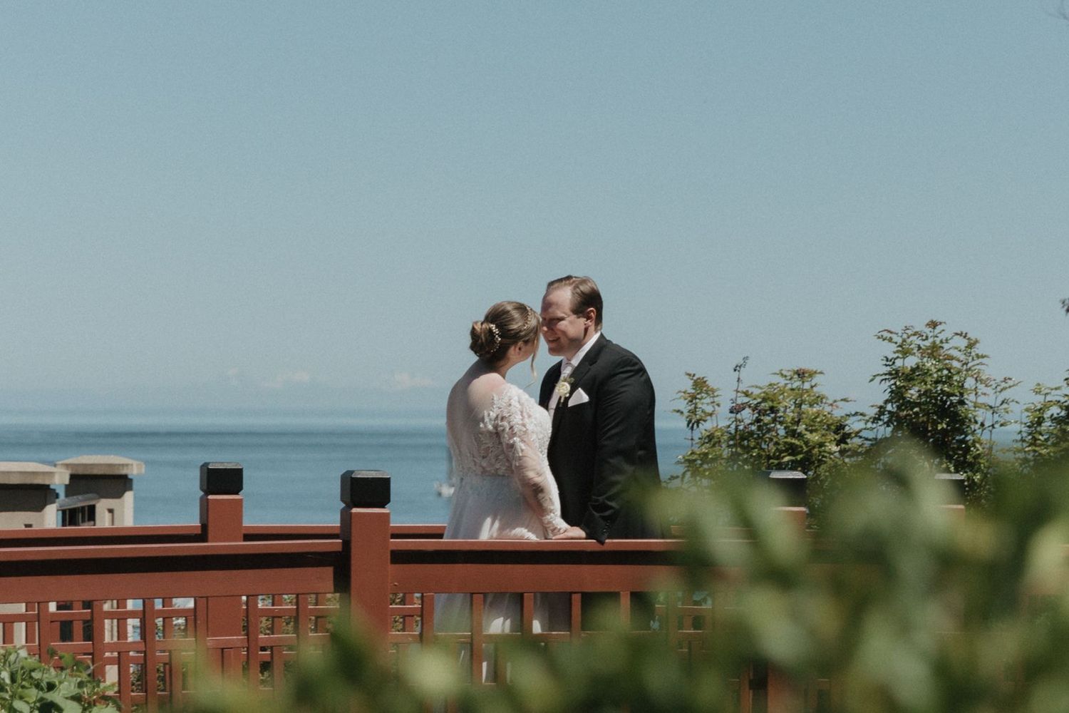 Bride and groom share a kiss on a red wooden bridge overlooking a serene blue ocean on their wedding day.