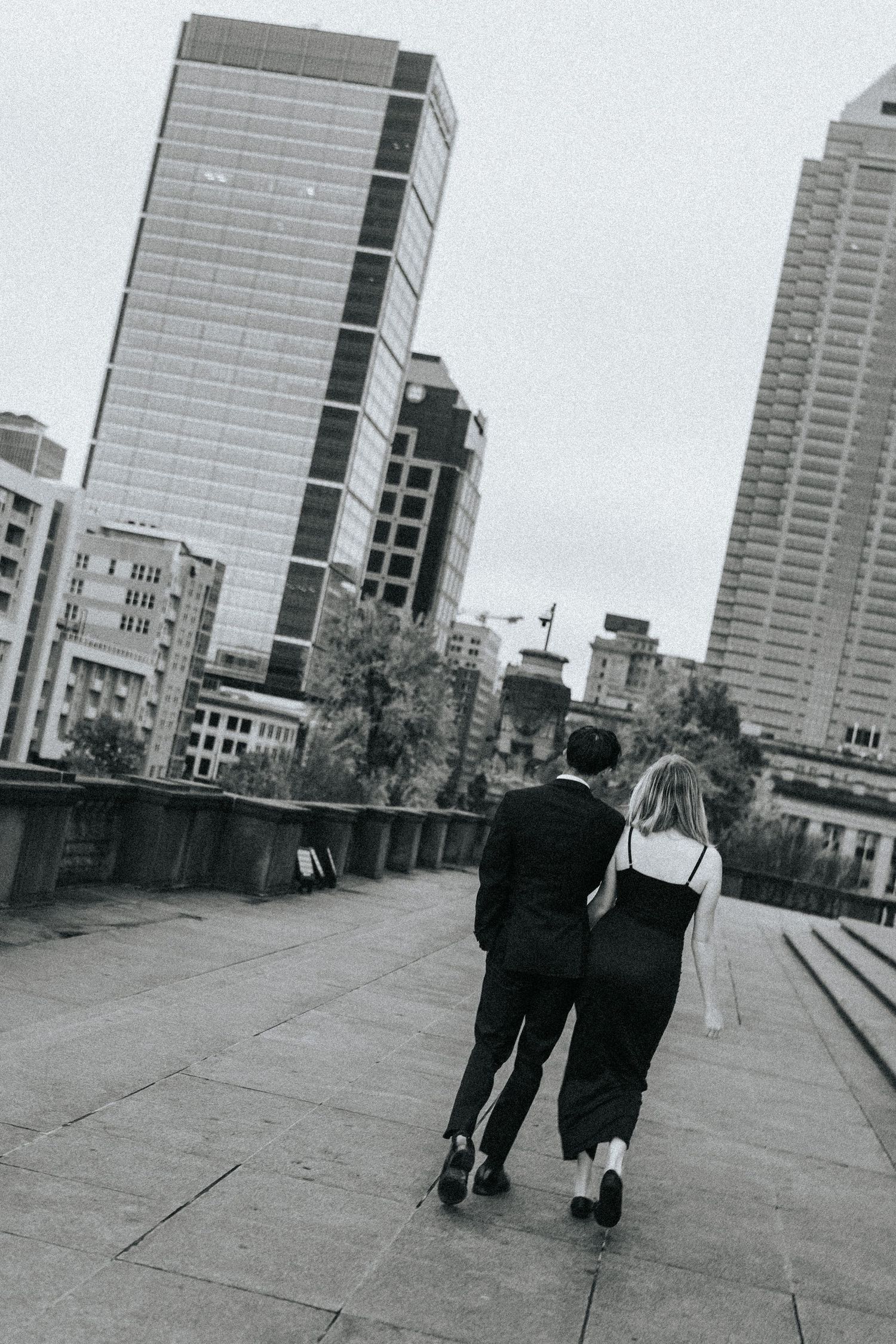 Black and white photo of two people walking along an urban rooftop with city buildings in background.