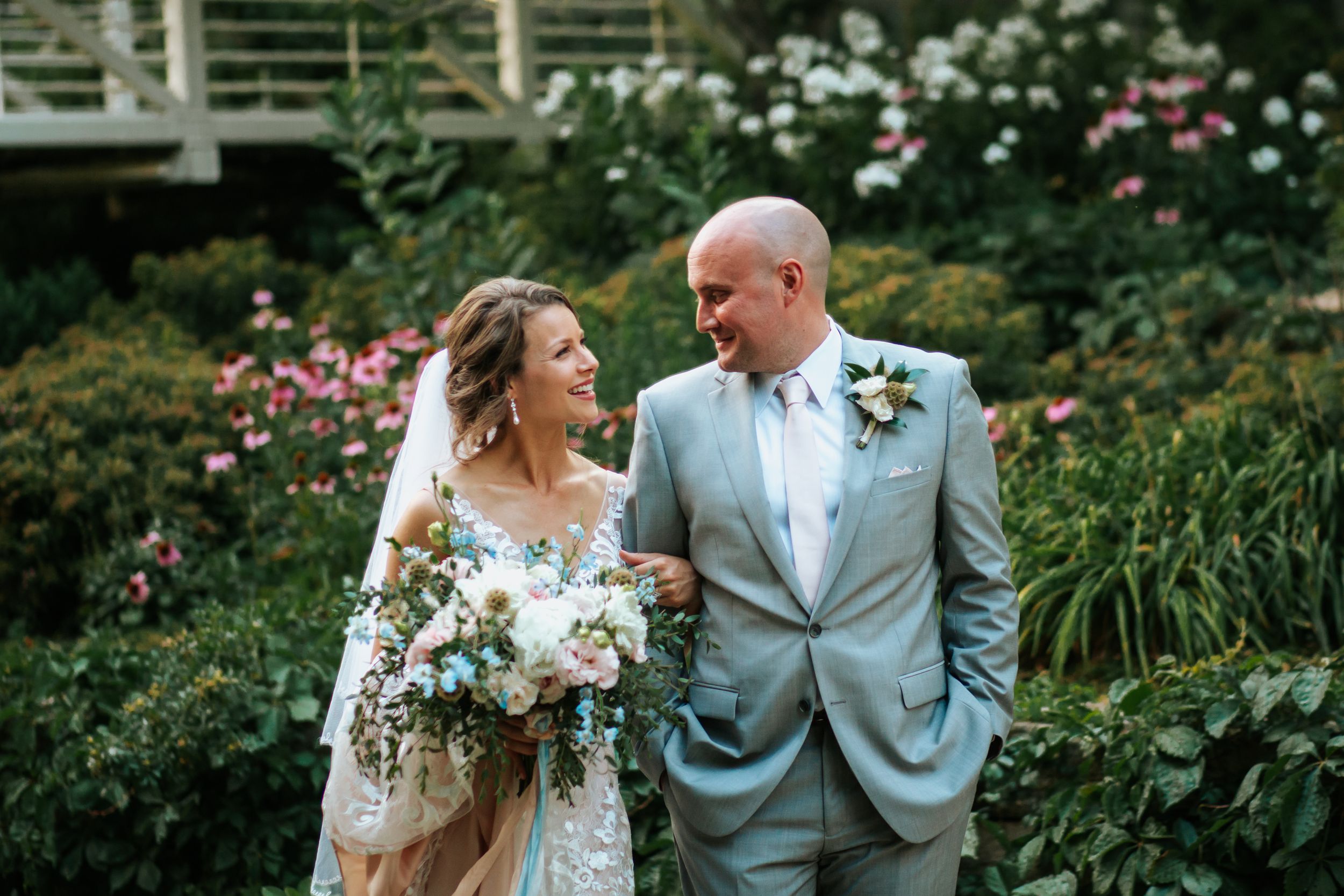 A romantic wedding couple poses in a lush garden setting with pink roses and greenery in the background.
