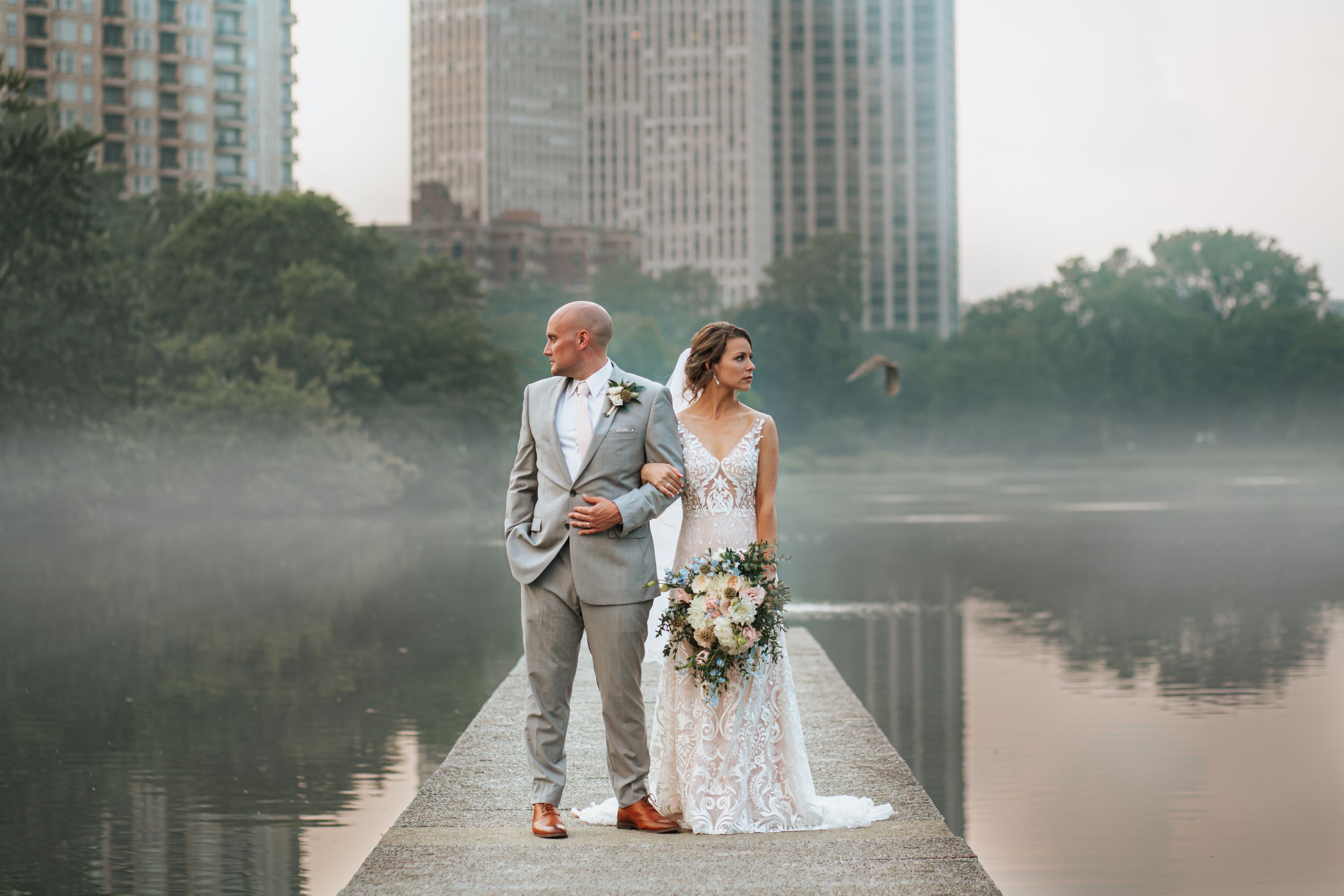 A couple in wedding attire stands on a misty dock overlooking a serene lake with city buildings in the background.