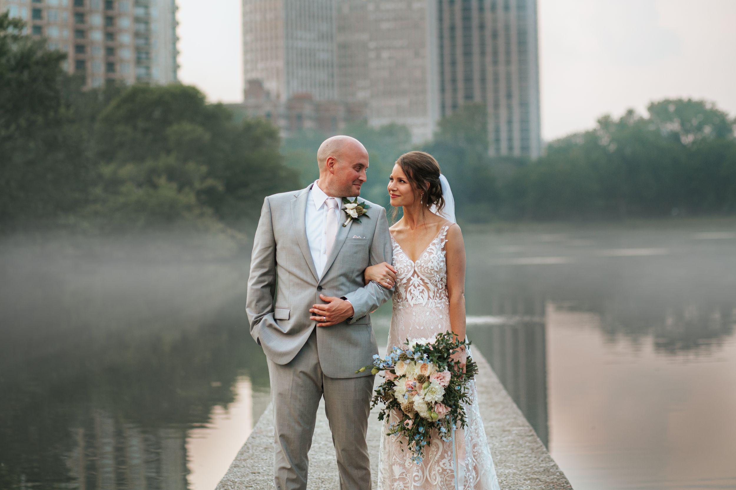 A couple in wedding attire stands by a misty lake with city buildings in the background.