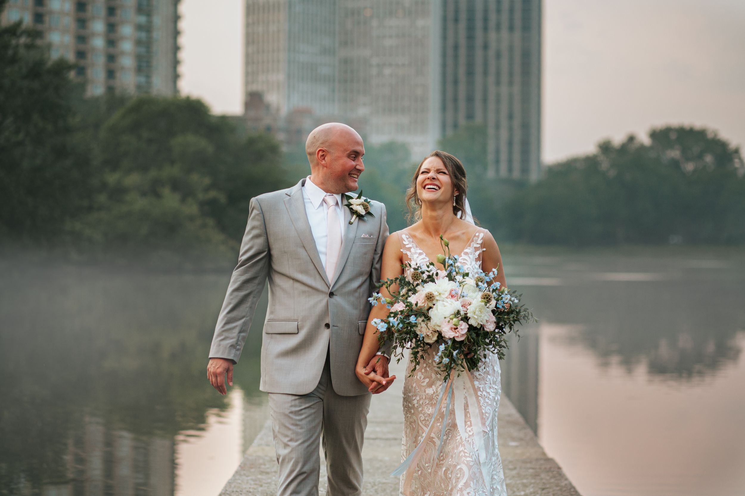 A couple in wedding attire walks together and shares romantic moments near a lake with city buildings in the background.