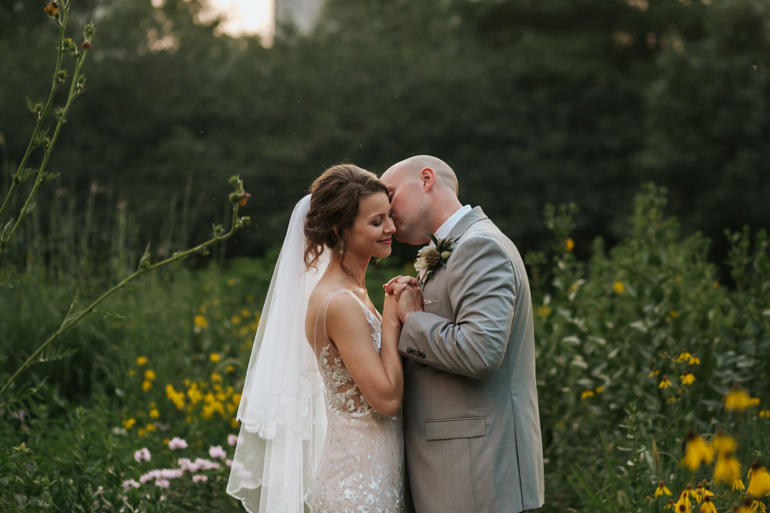 Bride and groom share romantic moments in a lush garden setting with yellow wildflowers at sunset.