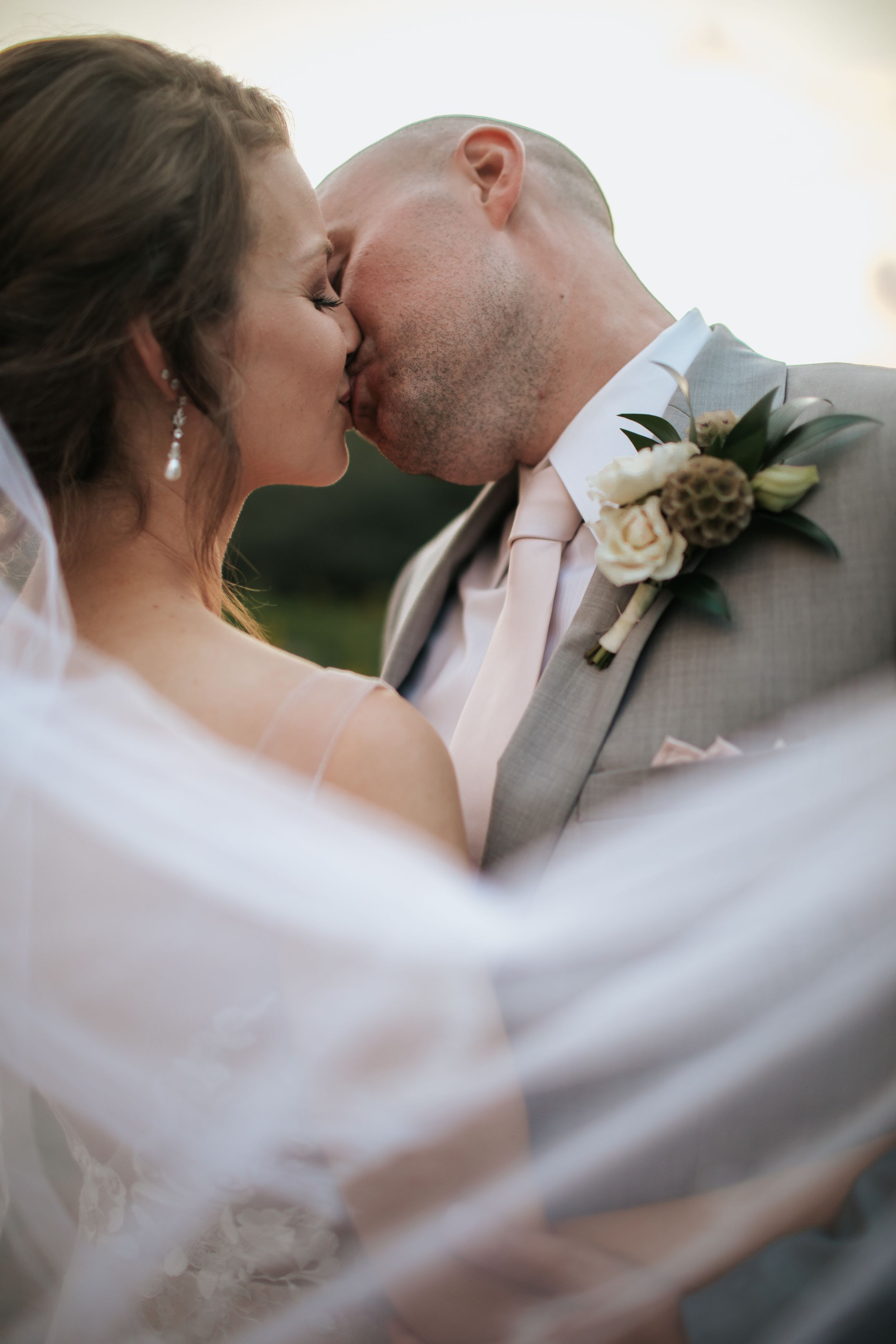 A romantic wedding kiss captured through flowing white veil with a boutonniere visible on the groom's lapel.