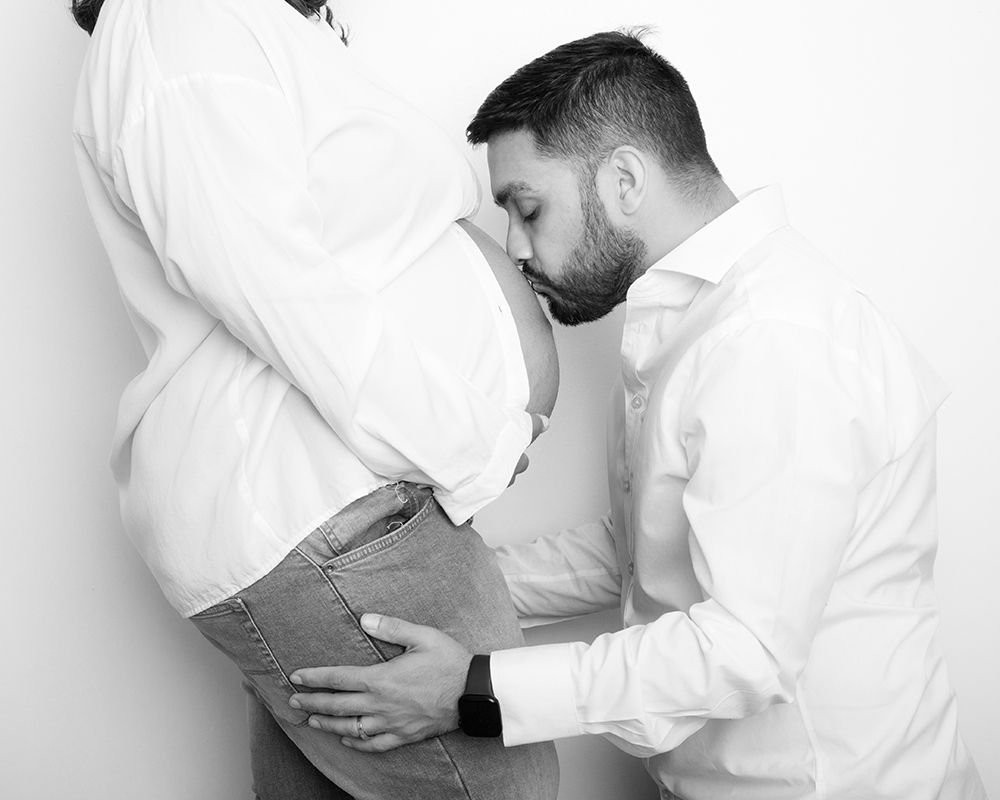 Black and white photograph of a man with a beard kissing a pregnant woman's baby bump while gently holding her hip. High Wycombe