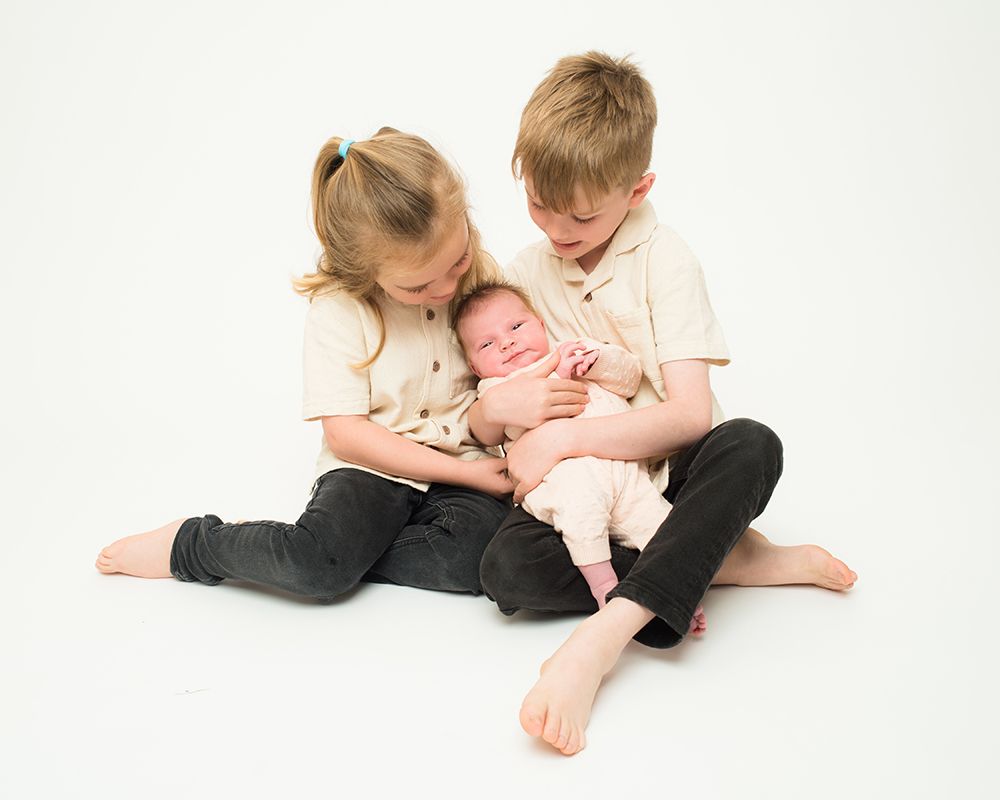 Two young children sitting on the floor holding a baby, all dressed in light-colored tops and dark pants, against a plain white background.