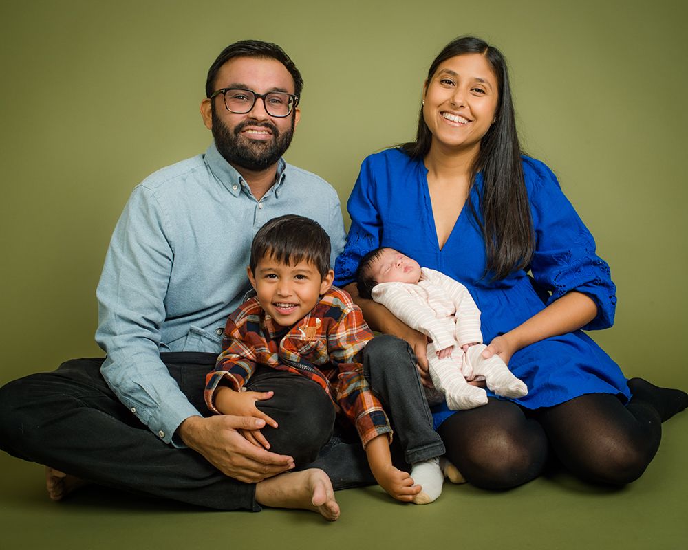 A smiling family of four, including parents, a young boy, and a newborn baby, sitting on a solid olive-green background. Abigail's Studio