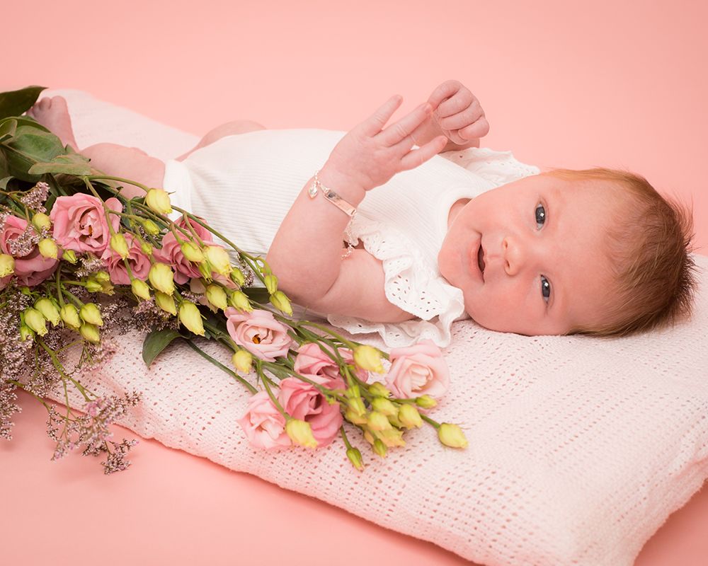 Newborn baby in a white outfit lies on a pink blanket with a bouquet of pink and yellow flowers beside them, set against a pink background.
