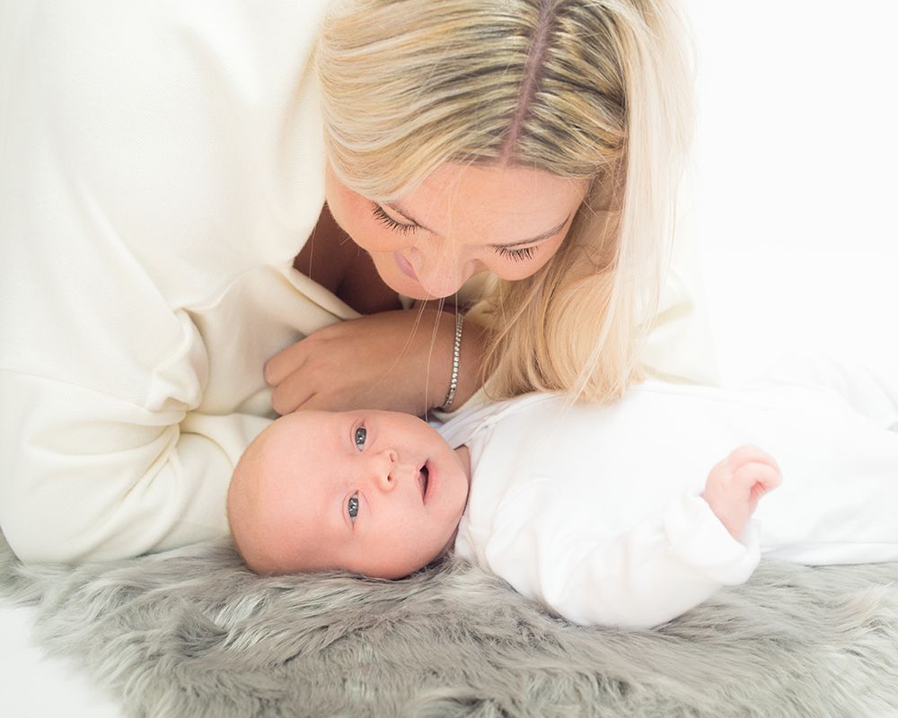 Blonde mother leans over her wide-eyed newborn baby dressed in white who rests on a faux fur rug. Photography Abigails Studio High Wycombe