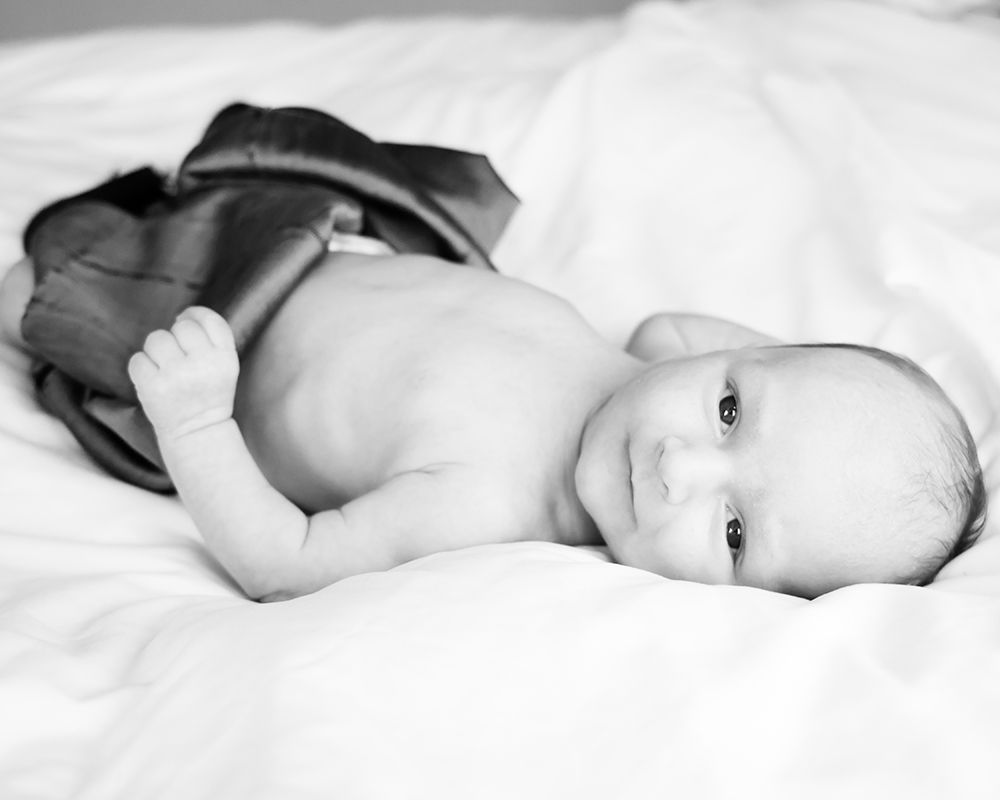 Black and white close-up of a bare-chested newborn baby lying on a white, rumpled sheet with dark fabric draped over the lower body.