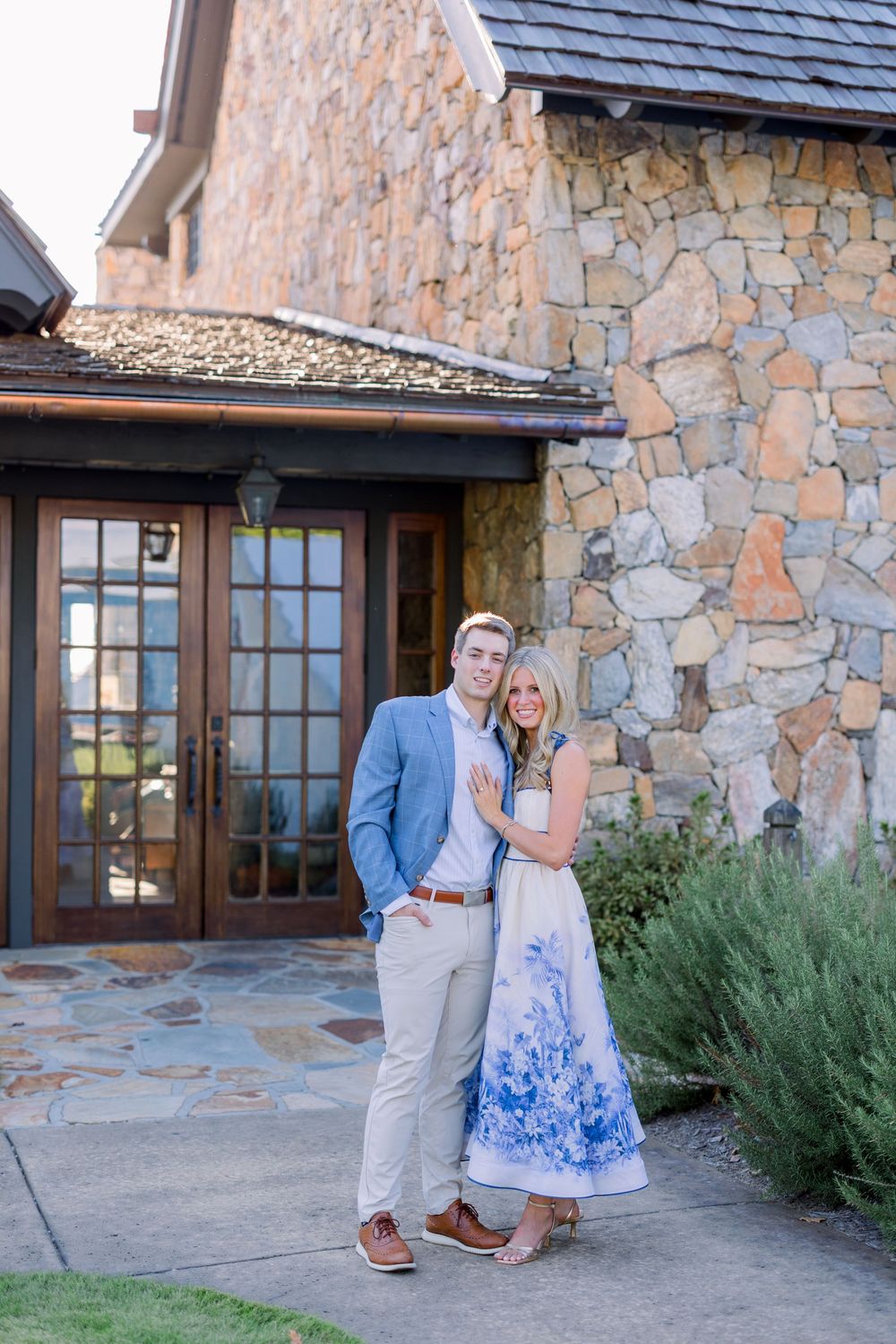 A couple stands together outside a rustic stone building with a slate roof and wooden doors.