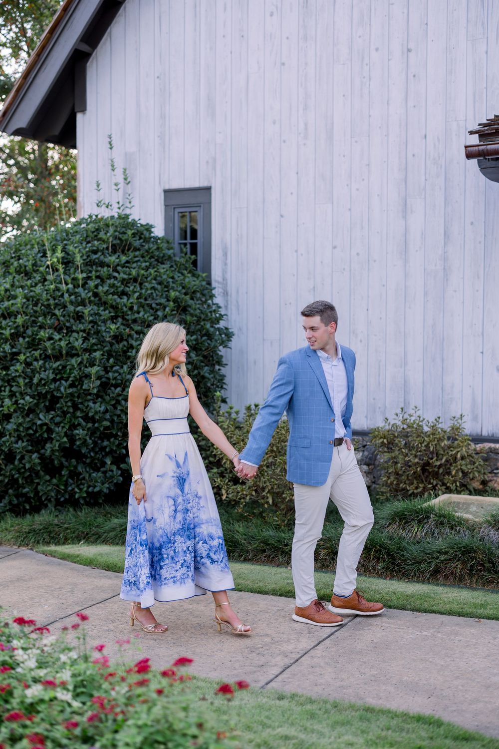A couple in summer attire holds hands by a white barn wall with blue floral dress and light blue blazer.