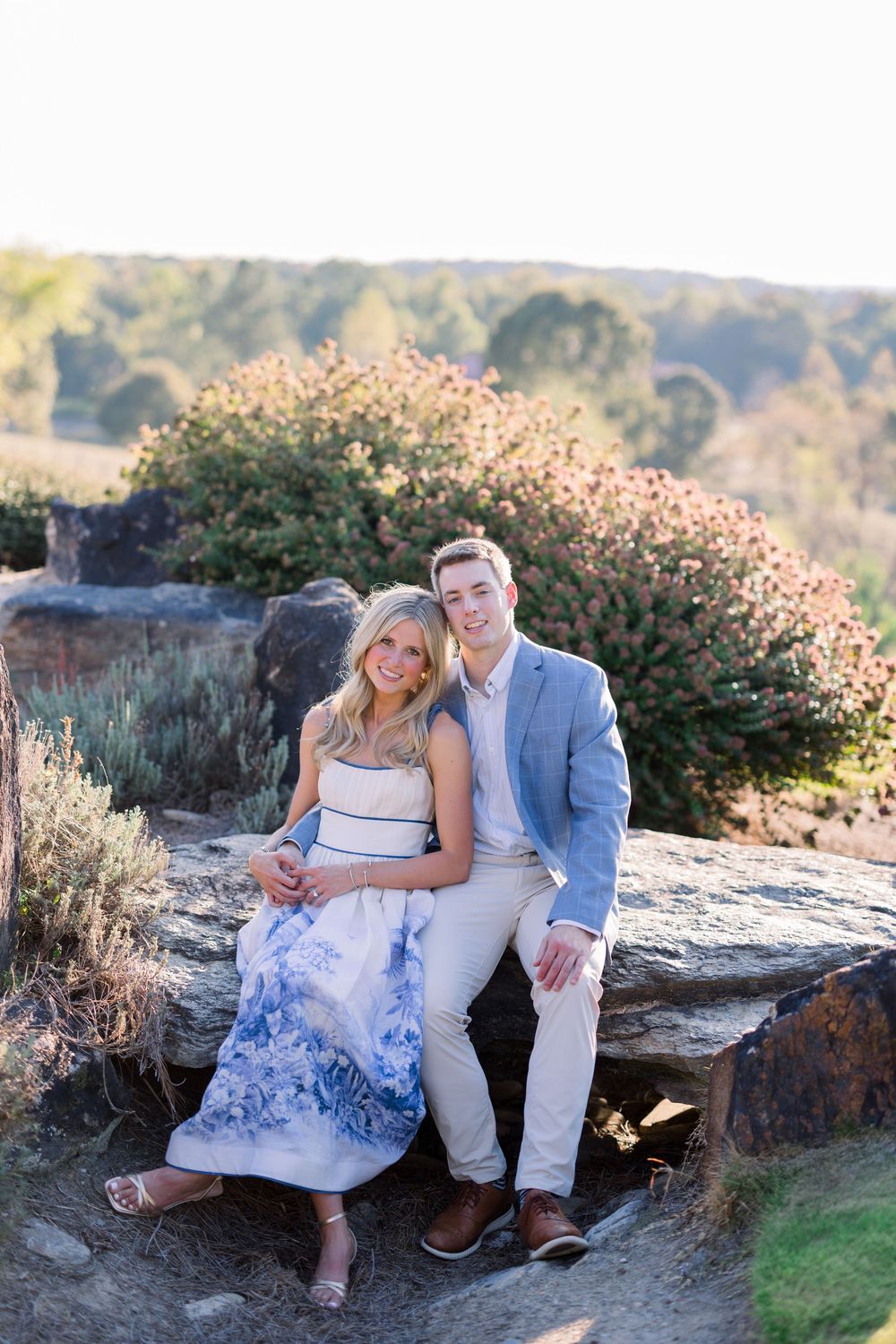 Couple shares intimate moments while sitting on natural stone ledge surrounded by greenery during engagement session.