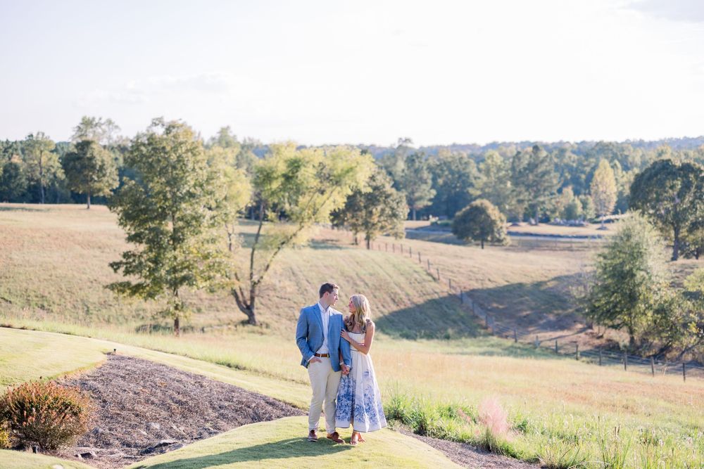 A couple walks along a scenic hilltop trail overlooking a rural landscape during a sunny autumn day.