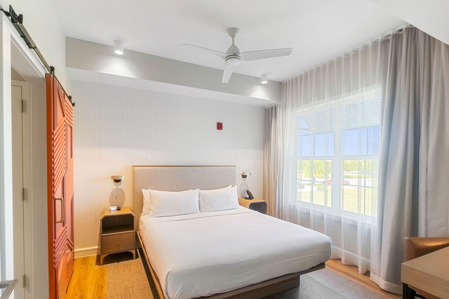 Modern hotel room interior with white bedding, wooden furnishings, and natural light streaming through sheer curtains.